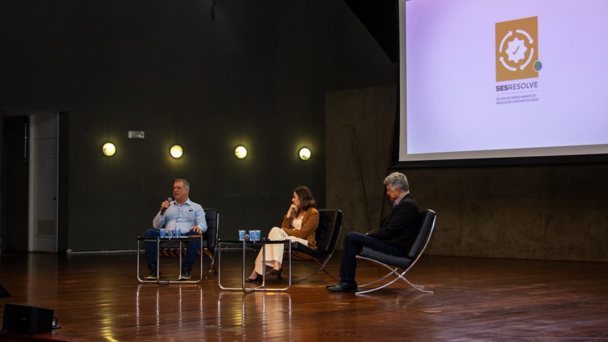 Três pessoas sentadas no palco durante o lançamento da plataforma SESResolve, com telão exibindo o logotipo do sistema ao fundo.