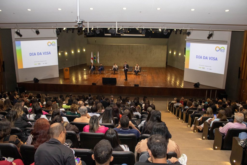 O público acompanha uma fala no palco durante o evento. Um dos palestrantes está de pé ao microfone, à direita, enquanto quatro homens permanecem sentados ao fundo. Os telões repetem a identidade do “Dia da Visa”, reforçando a ambientação institucional.