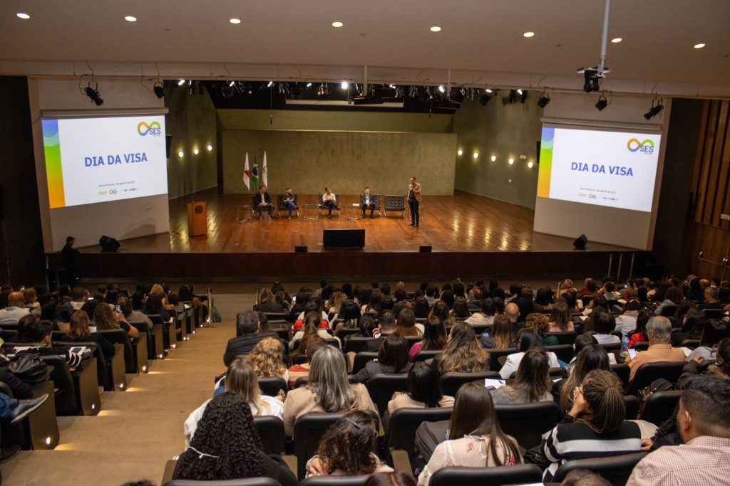Vista ampla do auditório lotado durante o evento “Dia da Visa”. No palco, cinco homens estão sentados e um deles está em pé, falando ao microfone. Dois telões laterais exibem a identidade visual do evento, com os dizeres “DIA DA VISA” e “Belo Horizonte, 7 de agosto de 2025”. Ao fundo, estão as bandeiras de Minas Gerais, Brasil e SES-MG.