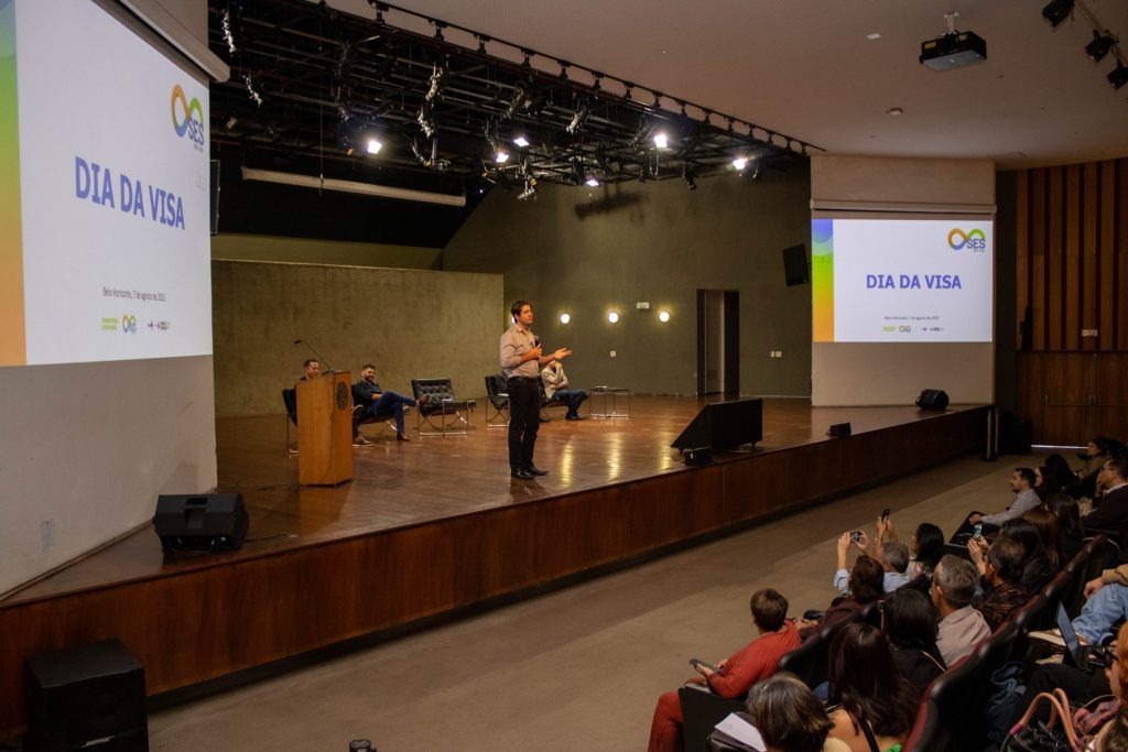 Vista lateral do auditório, com foco no secretário de Estado de Saúde, Fábio Baccheretti, em pé no palco durante seu pronunciamento. Ao fundo, os demais participantes estão sentados. O público aparece em primeiro plano, atento, e muitos usam o celular para registrar o momento.