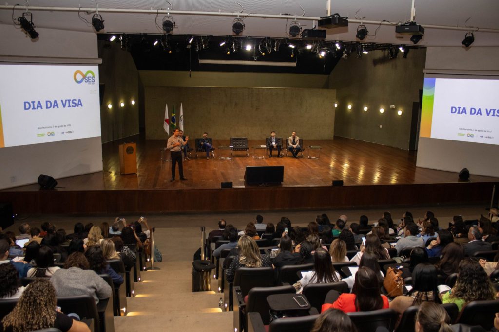 Perspectiva central do auditório, com Fábio Baccheretti falando ao microfone no centro do palco. Ao fundo, estão os demais convidados. O ambiente é bem iluminado, com destaque para a participação ativa do público, que acompanha a fala e faz registros com seus celulares.