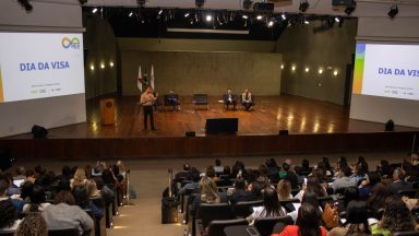 Perspectiva central do auditório, com Fábio Baccheretti falando ao microfone no centro do palco. Ao fundo, estão os demais convidados. O ambiente é bem iluminado, com destaque para a participação ativa do público, que acompanha a fala e faz registros com seus celulares.