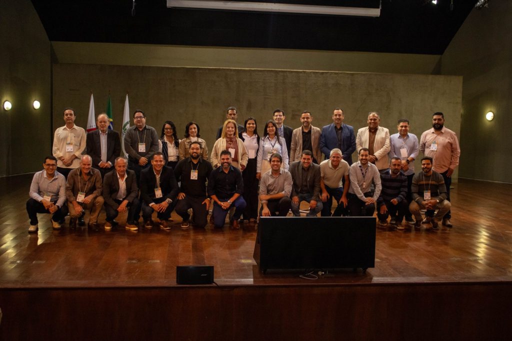 Foto oficial com os participantes do evento "Dia da Visa", reunidos no palco para o registro coletivo. O grupo é composto por cerca de 30 pessoas, entre representantes da SES-MG, consórcios intermunicipais e parceiros da Vigilância Sanitária. Ao fundo, estão as bandeiras oficiais.