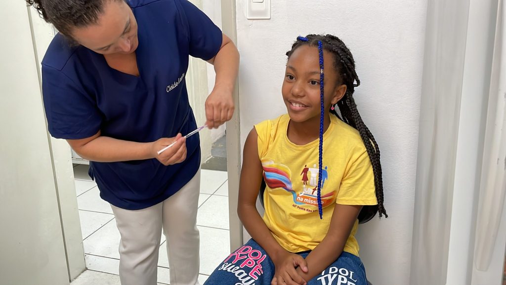 Preparação de vacina em unidade de saúde: profissional aplica dose em menina sorridente, sentada, com tranças coloridas.