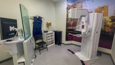 Sala de mamografia com equipamentos médicos. À direita, aparelho de mamografia com tela digital e bandeja de posicionamento. À esquerda, monitor e equipamento com tela de vidro. No centro, cadeira preta, avental de chumbo pendurado e suporte para filmes radiográficos. Ao fundo, mural decorativo com prédios e árvores.